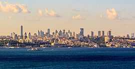 Skyline of a portion of the city, including several skyscrapers interspersed among low- and mid-rises, several historic buildings, parks and hilly terrain, and the waterway known as Bosphorus