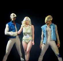 A blond woman in a nude colored leotard is flanked by two males in nude body suit and blue jacket. The woman's chest and the hand and head of the two males are covered by skeletal masks. The woman has glitter surrounding her eyes.