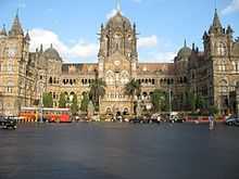 A brown building with clock towers, domes and pyramidal tops. Also a busiest railway station in India. A wide street in front of it