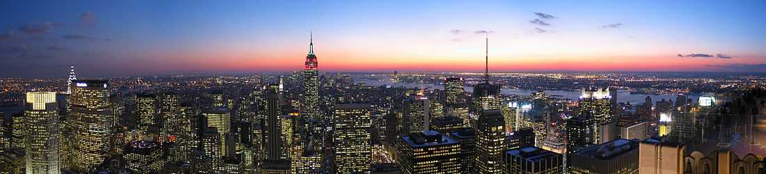 New York skyline at sunset, most of the blocky buildings with their windows lit up, and centered on a Empire State Building lit up red at its top.