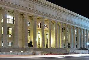 A long light-colored stone building seen from across the street at night. It has round fluted columns with ornate capitals running across its front, from one side of the image to the other. Behind them are large arches with windows. At the top is a wide frieze with "State Education Building" carved into the entablature.