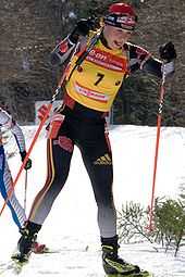 A woman cross country skis uphill towards the camera, holding a red ski pole in each hand. She wears black winter sportswear, a red cap and a yellow jersey with the number 7. A second skier behind her can be seen on the left.