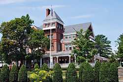 An ornate brick house with a pyramid-roofed tower on the front seen from slightly below. In front is a flagpole and some tall trees, with shrubbery and a chain link fence at the bottom of the image, closer to the camera.