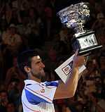 A brown-haired man in a white tennis shirt with light blue sections and red stripes with the trophy