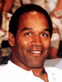 A black man smiling at a camera while wearing a white shirt.