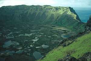 Colour photo from a cliff top down onto a pond strewn marsh backed by high cliffs, an area of sea is visible through a gap in the cliffs.