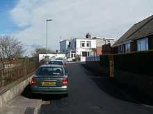 Under a partly cloudy sky, a concrete roadway recedes into the distance where a two-storey, flat-roofed, white stone building can be seen. A green car is parked behind a row of three cars. On the left is a low concrete wall with iron railings and a lamp post. On the right are a hedge and wall surrounding a bungalow.