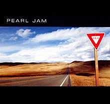 A triangular yield sign alongside a desertic road. Above and below the image are black bars, the top one having the text "Pearl Jam".