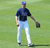 A man standing on a grassy surface wearing a baseball uniform with a dark blue shirt and white pants