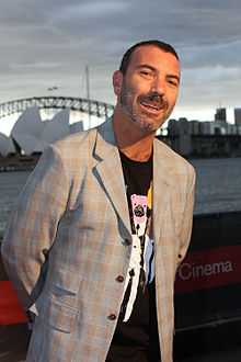 A 46-year-old man is shown in a three-quarter shot. He is leaning slightly to his left. He wears a peppery beard with very short hair and is smiling with his arms behind. His coat is checked grey and brown which obscures his black tee-shirt and its cartoon print. In the background is the Sydney Opera House and Harbour Bridge with Port Jackson behind him. The sky is overcast.