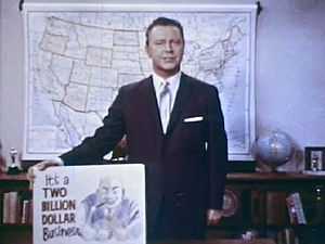 A still shot from the 1965 film "Perversion for Profit" showing a man in a suit holding a placard. The placard features a cartoonish image of a leering man and the phrase "It's a two billion dollar business"