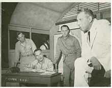 In a "war room", three military men watch a worried man in civilian dress