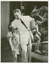 Pinza, in a white suit, walks along holding a mixed-race boy, about 7 or 8 years old, while listening to a slightly older girl who walks next to him.