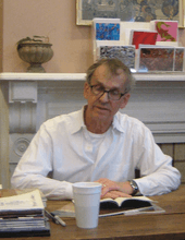  grey haired man in white shirt with glasses seated at table reading from a book