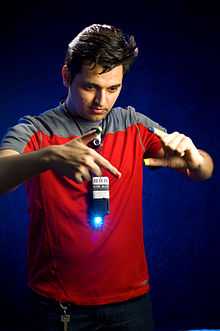 A man holds his hands in front of cellphone-sized pendant with a shining blue light. His fingertips are painted in different colors.