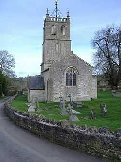 Gray building with arched windows. Square tower surmounted by a weather vane. Gravestones and crosses in grass in the foreground seperated from the road by a stone wall.