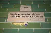 A green brick wall with a white sign reading "Wer die Vergangenheit nicht kennt, / ist dazu verurteilt, sie zu wiederholden. / (G. Santayana 1863–1953, Philosoph)