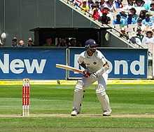 An Indian cricketer in a blue helmet, waiting to receive a delivery. He is standing in front of his wicket, with spectators in a stand behind him.