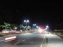 A wide road with lane markings, beautified median and decorative street lights