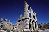 Ruins of a three-story masonry building rise into a cloudless, dark blue sky. The building is roofless, and large sections of it walls are missing. Masonry rubble lies about the building, which has openings for doors and windows but no glass or wood. Another ruin is nearby, and barren hills are visible in the distance.