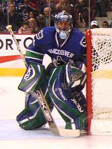 A masked ice hockey goaltender wearing a blue jersey with blue and green pads slightly crouched looking forward.