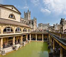 Photograph of the Baths showing a rectangular area of greenish water surrounded by yellow stone buildings with pillars. In the background is the tower of the abbey.