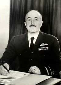 Half-length portrait of moustacioed man in military uniform with pilot's wings on left breast pocket, seated at desk with pen and papers