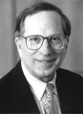 A man wearing glasses with a tie and a suite jacket looks to the camera and smiles