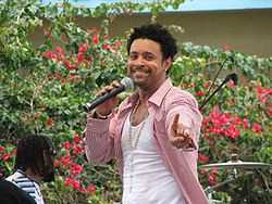 A man holding a microphone with one hard and extending his index finger with the other; he is wearing a white undershirt, red- and white-striped dress shirt and jewelry accessories. In the background is a man and vegetation.