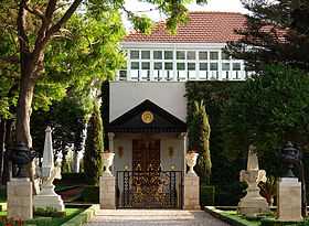 A view from the front of the Shrine of Bahá'u'lláh. The white building has a red tiled ceiling and an ornate door which includes inscriptions of the Bahá'í Most Great Name in modified Persian script. Thick vegetation grows on all sides of the entrance.