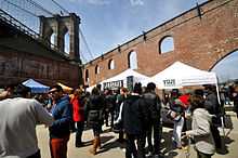 People crowd around white tents in the foreground next to a red brick wall with arched windows. Above and to the left is a towering stone bride.