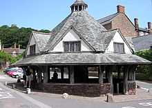 Street scene with houses and shops on the left and an octagonal structure has a central stone pier which supports a heavy timber framework which carries a slate roof with central wooden lantern surmounted by a weather vane.