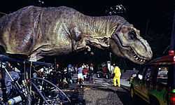 A life-sized Tyrannosaurus robotic model, with hydraulics  where the dinosaur's feet would be, touches a car in a movie set.