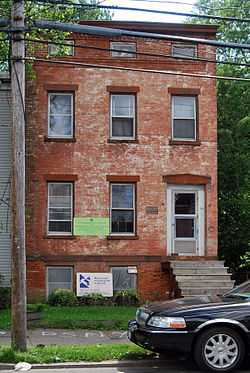 The front of a two-and-a-half story brick building with an exposed basement, seen from a nearby street, partially obstructed by the front of a parked car