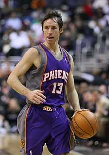 A basketball player stares to his right as he moves up the court with the ball. He is in a blue uniform with red and grey trim, and the words "Phoenix 13" on his chest.