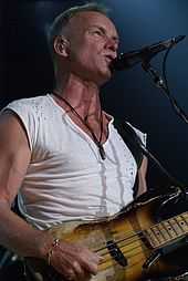 A man in a white shirt standing behind a microphone stand and holding a guitar