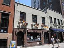 A two-story building with brick on the first floor, with two arched doorways, and gray stucco on the second floor off of which hang numerous rainbow flags.