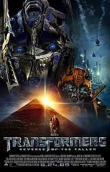 The faces of two robots stand atop a pyramid. A helicopter flies over an industrial facility on the right side of the image, and a young couple is seen in front of the pyramid. The film title and credits are on the bottom of the poster.