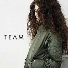 Lorde looking down with her face partially obscured by long curly dark hair, wearing a khaki-coloured bomber jacket and the text "TEAM".