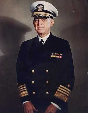 Man in dark blue suit and tie, wearing peaked cap and two rows of ribbons.