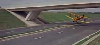 A yellow biplane flies underneath a deserted motorway bridge.