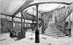 View of a wide branching staircase, leading off to the left and right top of the scene. Elaborate balustrades line the steps, down which a woman is walking. At the head of the stairs a wall clock is visible, and above that a segmented dome. A man and a woman sit in chairs in the foreground.