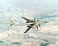 Top perpendicular starboard view of four-engine propeller-driven aircraft in-flight. The pale gray/white aircraft is flying above almost white terrain.