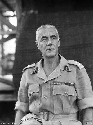 Grey haired man in Army shirt with sleeves rolled up. He is wearing rank badges and ribbons but no tie or hat.