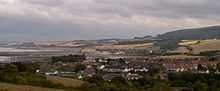 View of multiple houses with sea on the left and hills in the background.