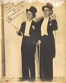 Dressed in tuxedos and top hats, Bert Wheeler (left) and Robert Woolsey (right) stand and smile for a publicity photo. An inscription appears in the top left corner.