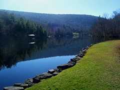 A small lake with a smooth surface reflecting the blue sky above and a hill beyond it with some buildings along the far shore. In the foreground, at lower right, the edge is marked by a stone retaining wall