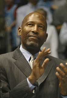 A man, wearing a gray suit, a shirt and a tie, is clapping his hands while looking up.