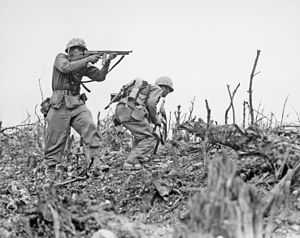 black & white photograph of two Marines advancing up a hill, the one on the left is firing an M1 submachinegun while the one on thr right dashes for cover