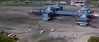 A massive, blue-grey spacecraft, roughly shaped like a plane but composed of multiple components, is on a runway surrounded by vehicles and buildings at the point of take-off.